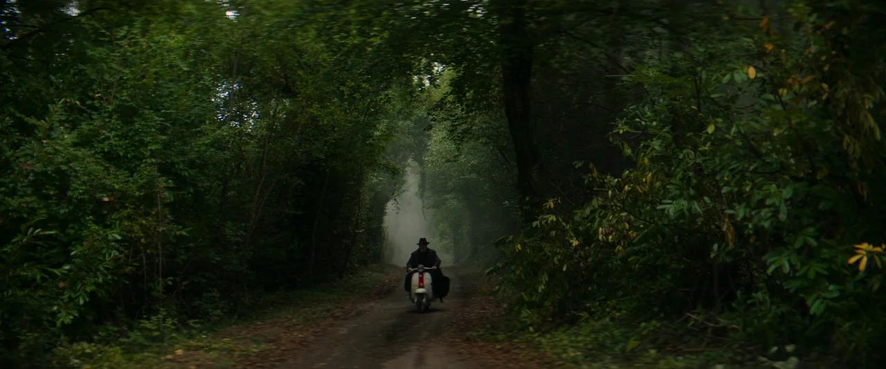The Pope’s Exorcist (2023) movie still: wide — A narrow dirt path cuts through a dense tunnel of overgrown lush green trees. A man in a hat rides a…