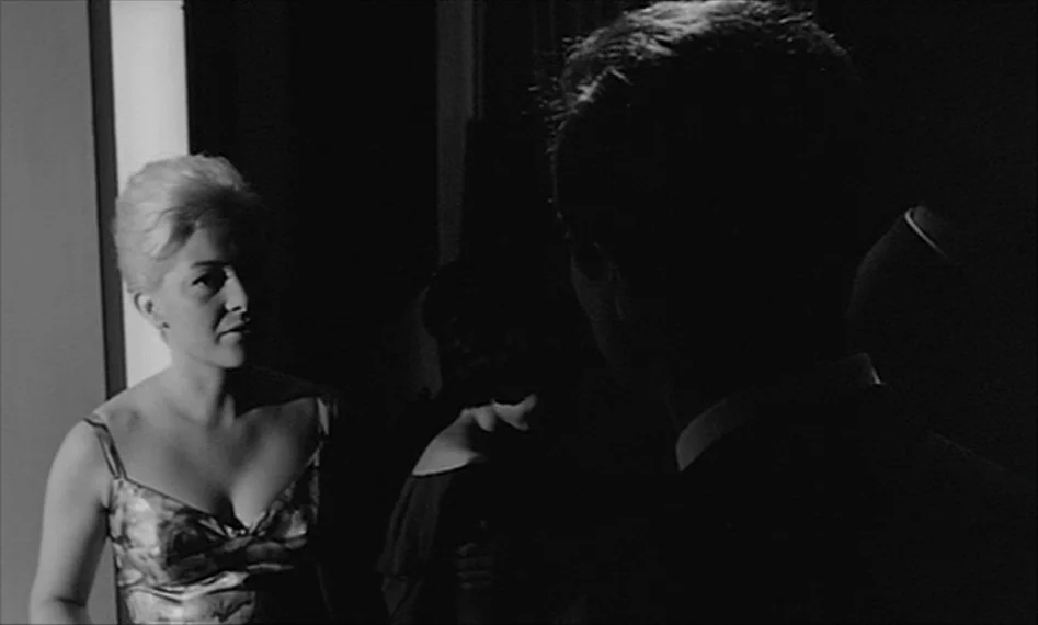 La Notte (1961) movie still: medium — A darkened interior hallway features a woman standing in a sliver of light. She looks at an obscured man whose…