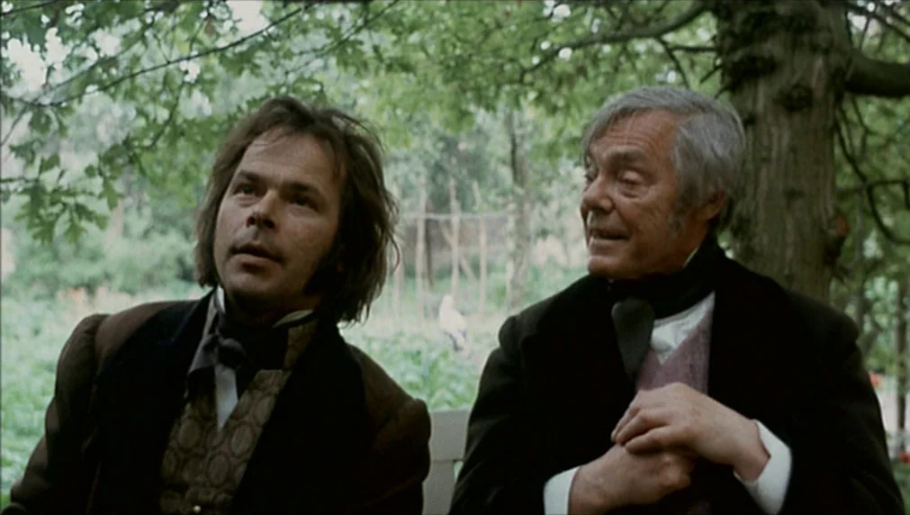 The Enigma Of Kaspar Hauser (1974) movie still: medium — Two men sit under the canopy of mature deciduous trees in a green space. They both look in…