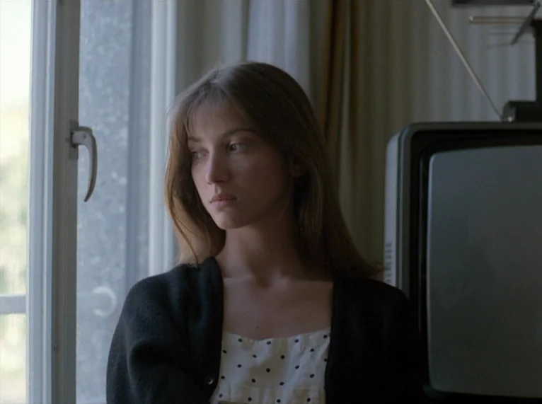 Hail Mary (1985) movie still: medium — A young woman stands near a window inside a living room with an old television. She gazes out the window…