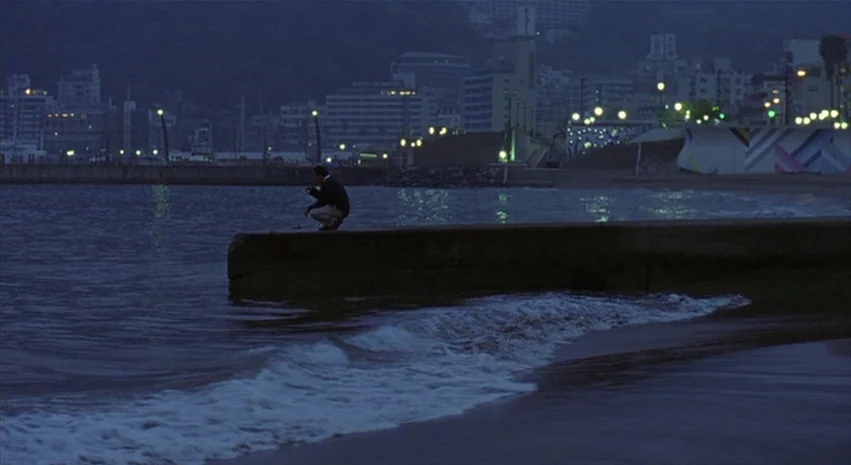 Yi-Yi (A One and A Two) (2000) movie still: wide — A man crouches on the edge of a concrete breakwater structure at night. The background shows a…