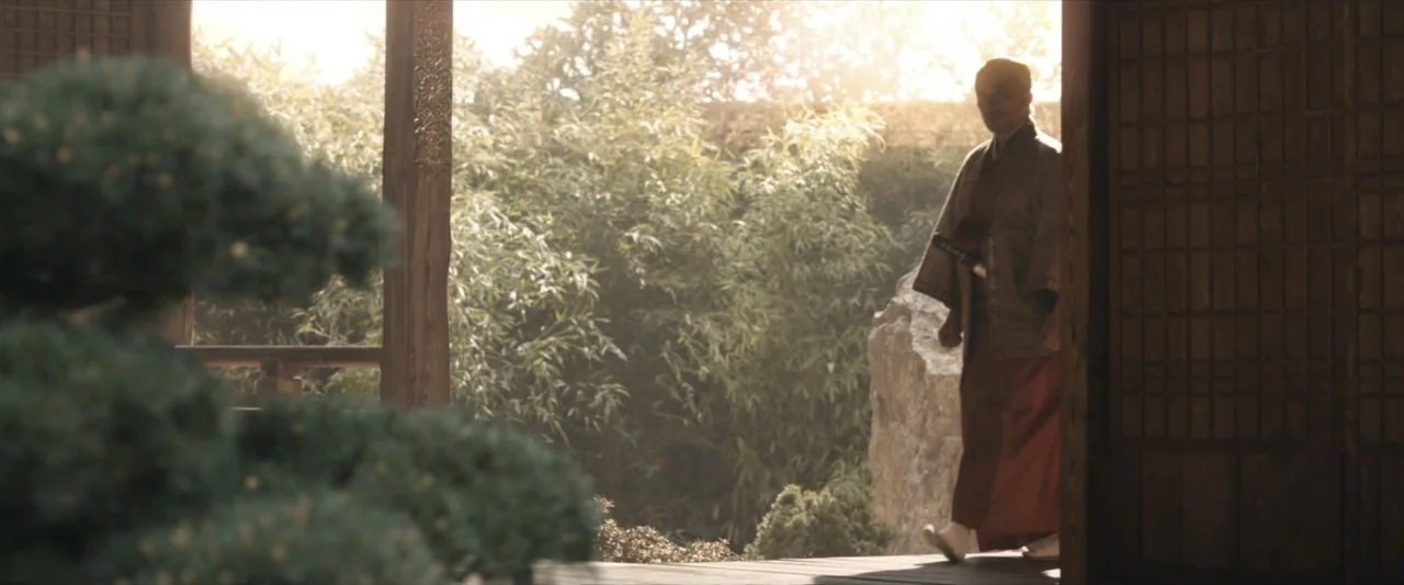 47 Ronin (2013) movie still: fullbody — A wooden Japanese veranda overlooks a garden filled with lush green bamboo and rock. A man in traditional…