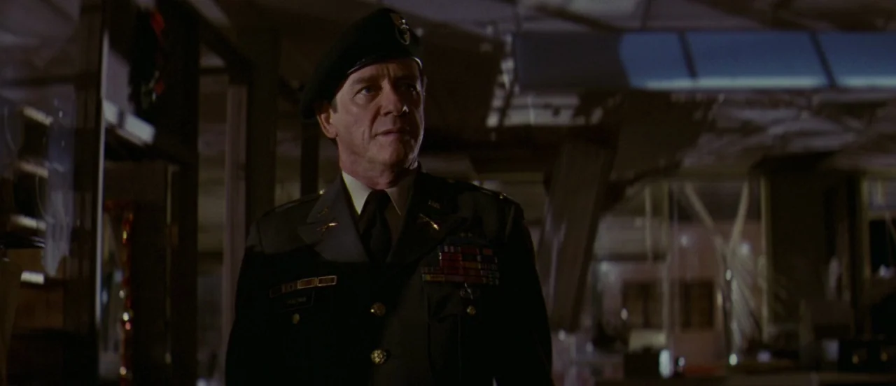 First Blood (1982) movie still: medium — A dimly lit room containing military furniture and cluttered office equipment in the background. A man in a…