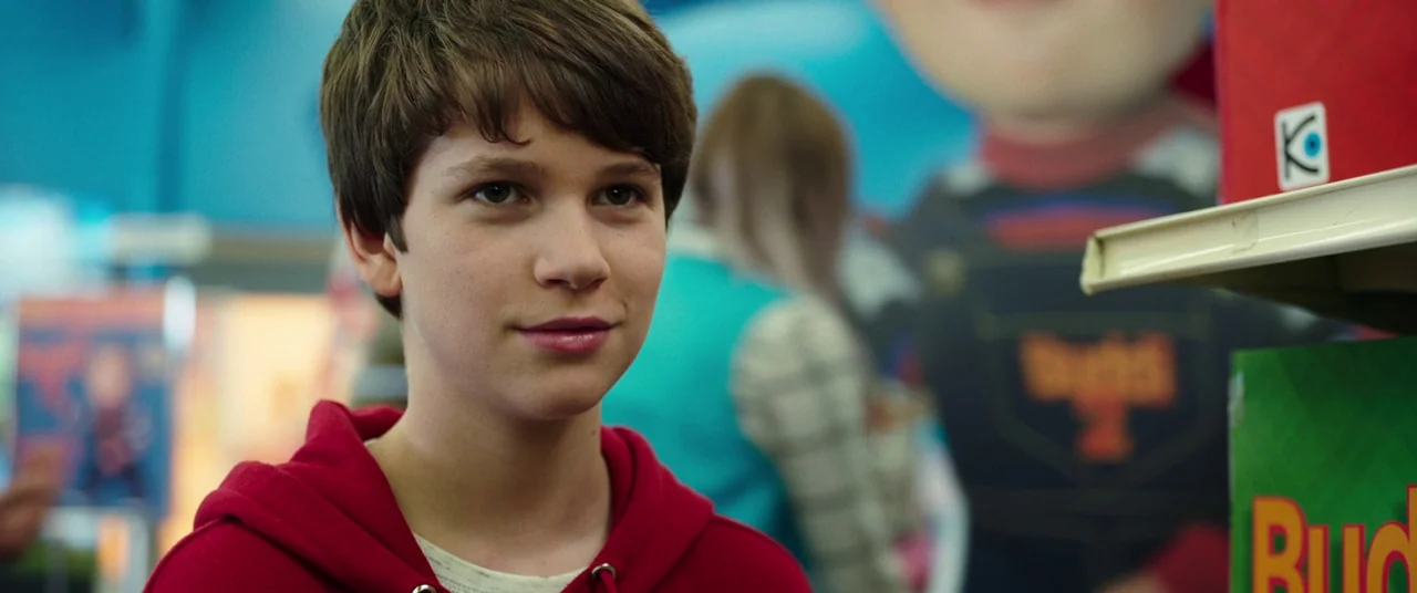 Child's Play (2019) movie still: closeup — A well-lit retail aisle is filled with colorful product packaging and displays. A young boy in a red…