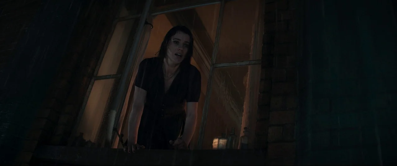Evil Dead Rise (2023) movie still: medium — A woman leans out of a dark, rain-streaked window at night. The surrounding wall is built of dark brick…