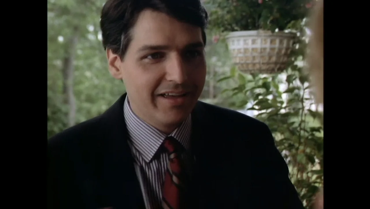 The Awakening (1995) movie still: medium — A man stands on a leafy porch with a hanging plant in background. He speaks to an off-screen person while…