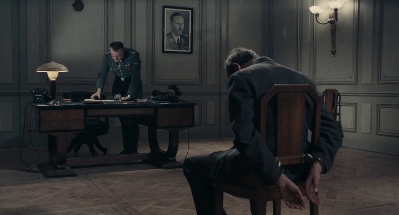 The Army of Shadows (1969) movie still: medium — A spartan room features gray paneled walls with a framed portrait of a man. A uniformed officer…