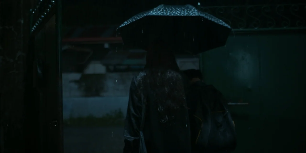 Nokturno (2024) movie still: medium — Two individuals stand in the rain at night beneath a large black umbrella. They face a closed metal gate…