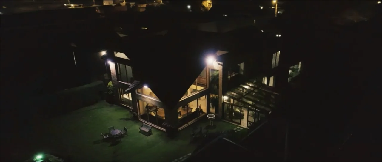 Loving is Losing (2019) movie still: establishing — A two-story suburban residence sits on a dark grassy lawn at night. The interior is brightly lit…