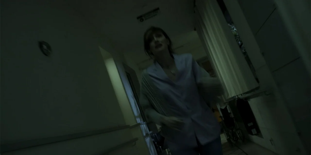 La coda del diavolo (2024) movie still: medium — A dark hospital corridor features white walls and a distant doorway under low light. A woman runs…