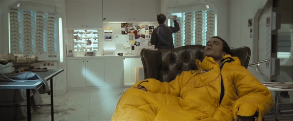 Moon (2009) movie still: medium — A white sterile living area contains a leather chair and a desk with posters. A man in a yellow sleeping bag rests…