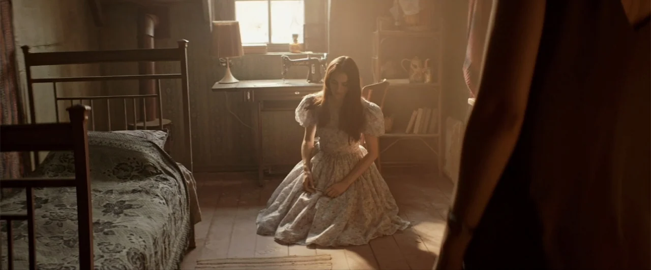 La herencia (2010) movie still: medium — A dimly lit room contains a wooden bed frame, a small desk, and shelving. A young woman in a floral dress…