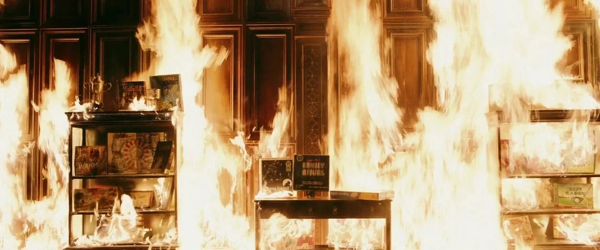 Ready or Not (2019) movie still: wide — A wood-paneled room is consumed by a massive, uncontrolled fire across the wall. Multiple shelves containing…