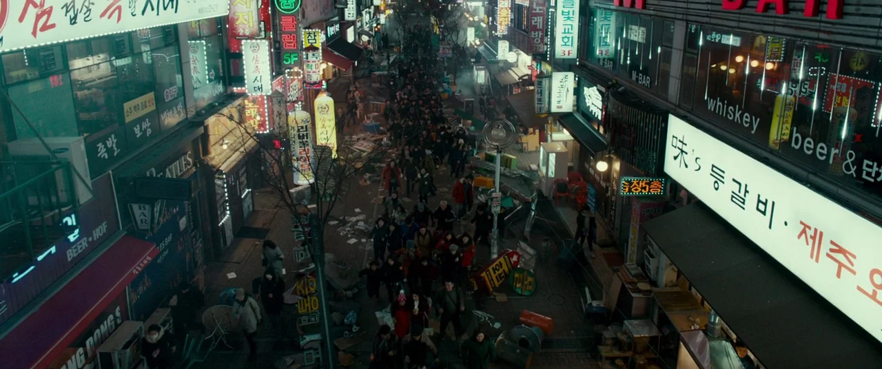 Colossal (2016) movie still: establishing — A dense urban alleyway lined with multi-story storefronts adorned with glowing neon signage in Korean. A…
