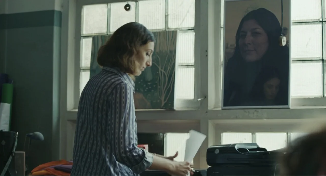 Belén (2025) movie still: medium — A workspace with glass blocks windows and artwork displayed on the ledge. A woman in a striped shirt stands near…