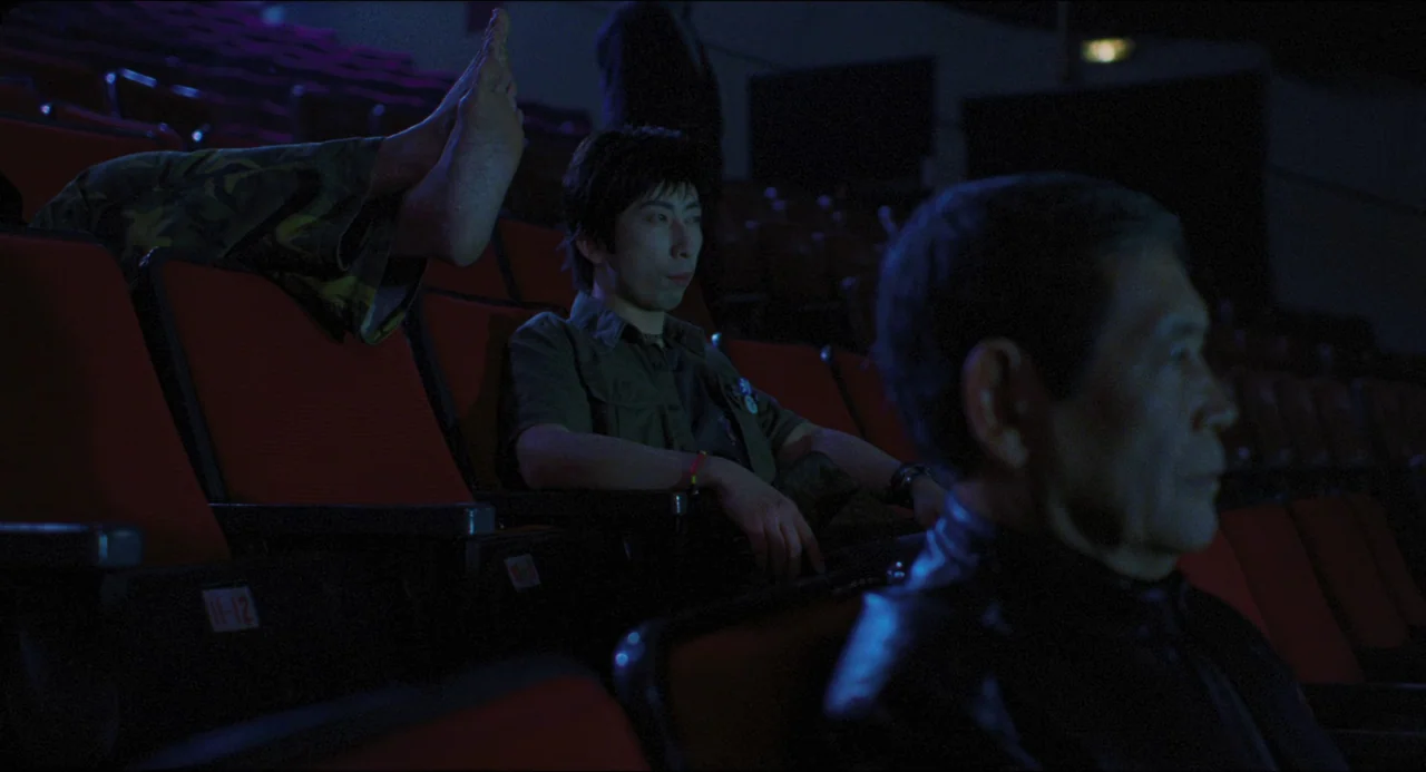 Goodbye, Dragon Inn (2003) movie still: medium — Rows of empty red seats fill a dark, quiet, indoor movie theater auditorium. A young man and an…