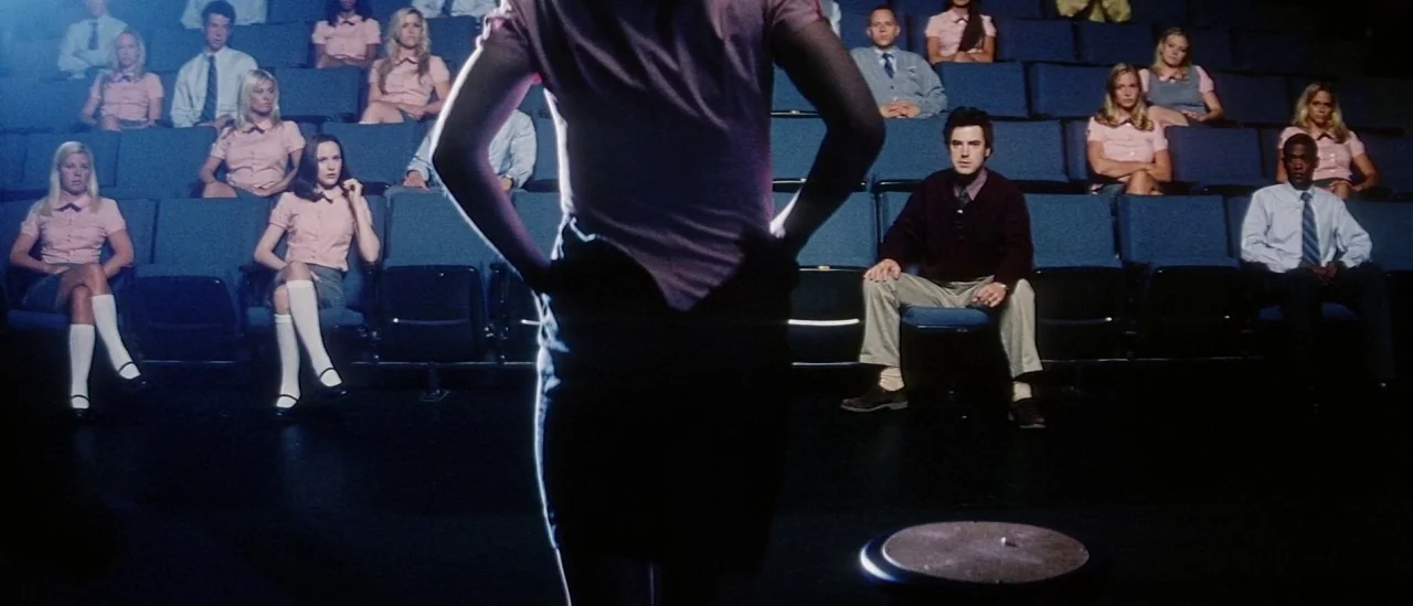 Pretty Persuasion (2005) movie still: medium — A dark auditorium is filled with rows of blue seats and many people. A woman stands in the foreground…
