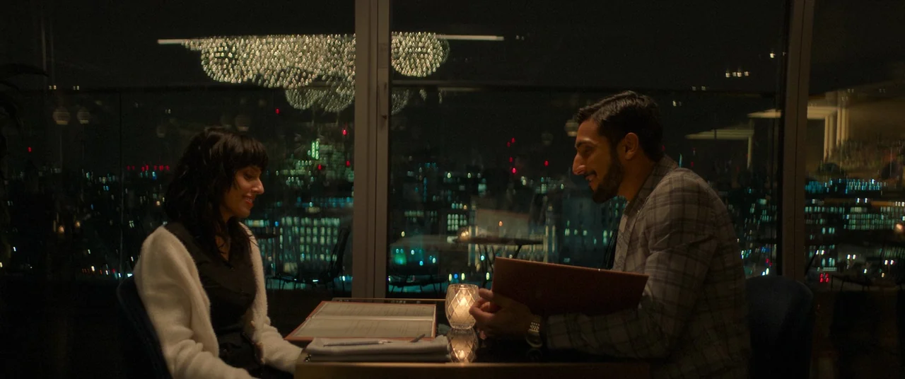 Polite Society (2023) movie still: medium — A high-rise restaurant at night with floor-to-ceiling windows overlooking a blurred city skyline. A man…