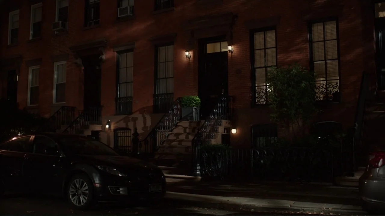 The Intern (2015) movie still: wide — A quiet residential street lined with red brick brownstone buildings at nighttime. A parked black sedan sits…
