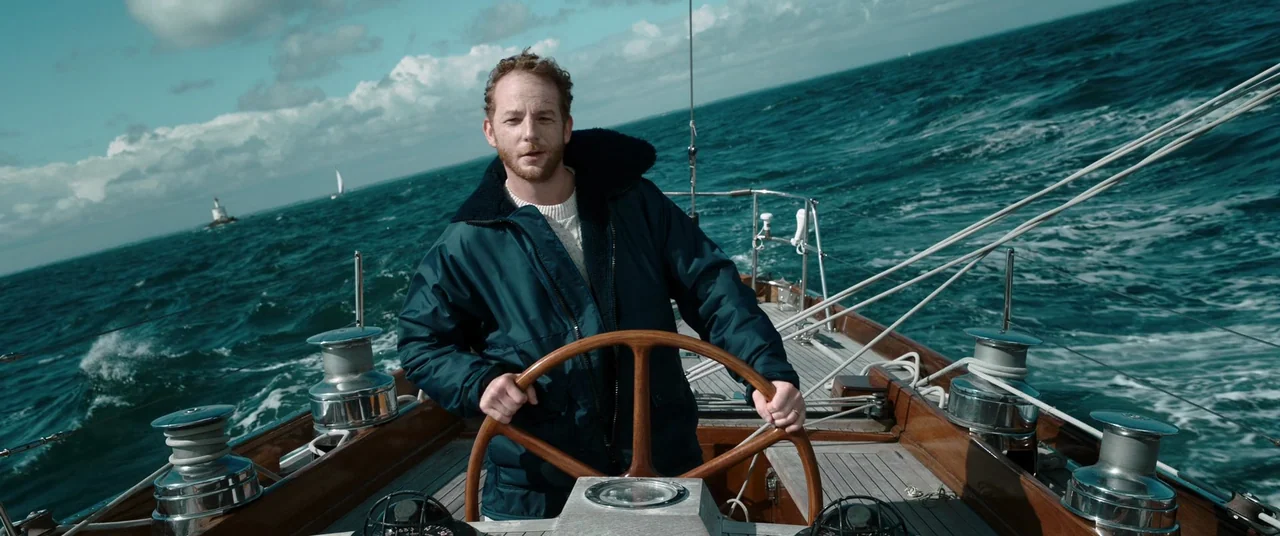 Oxygen (2021) movie still: medium — A wooden sailboat deck glides through choppy dark blue ocean water under a cloudy sky. A man holds onto the…