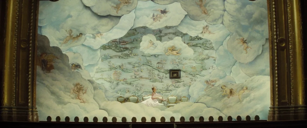 Anna Karenina (2012) movie still: wide — A theatrical stage with a large painted backdrop depicting clouds and cherubs. A lone woman sits on a small…