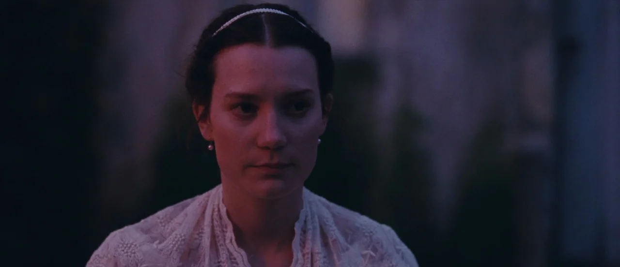 Madame Bovary movie still: closeup — A dark outdoor space with indistinct textures visible in the shadowed background. A young woman in a lace…