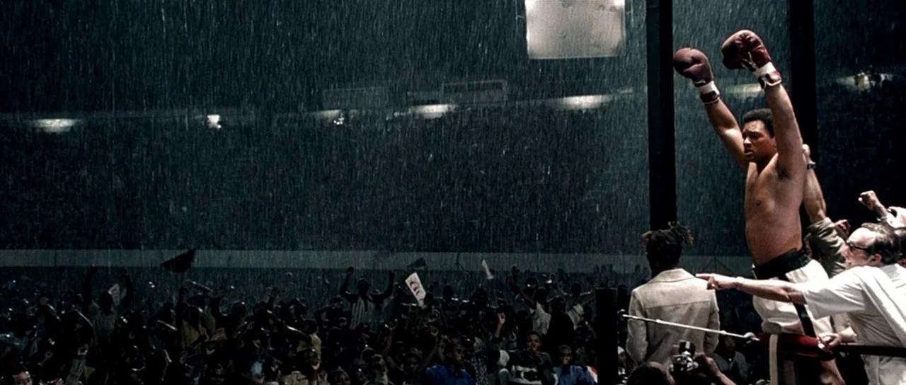 Ali (2001) movie still: wide — A vast open-air stadium during a heavy downpour filled with a cheering crowd. A boxer in trunks and red gloves stands…