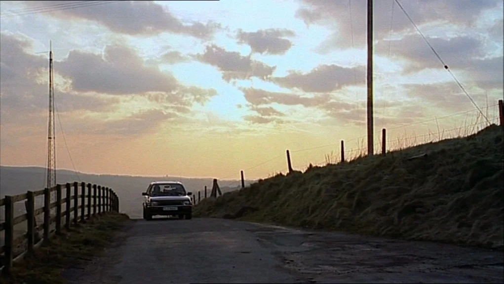 24 Hour Party People movie still: wide — A rural asphalt road cuts through a grassy hillside under a sunset sky. A single car travels down the lane…
