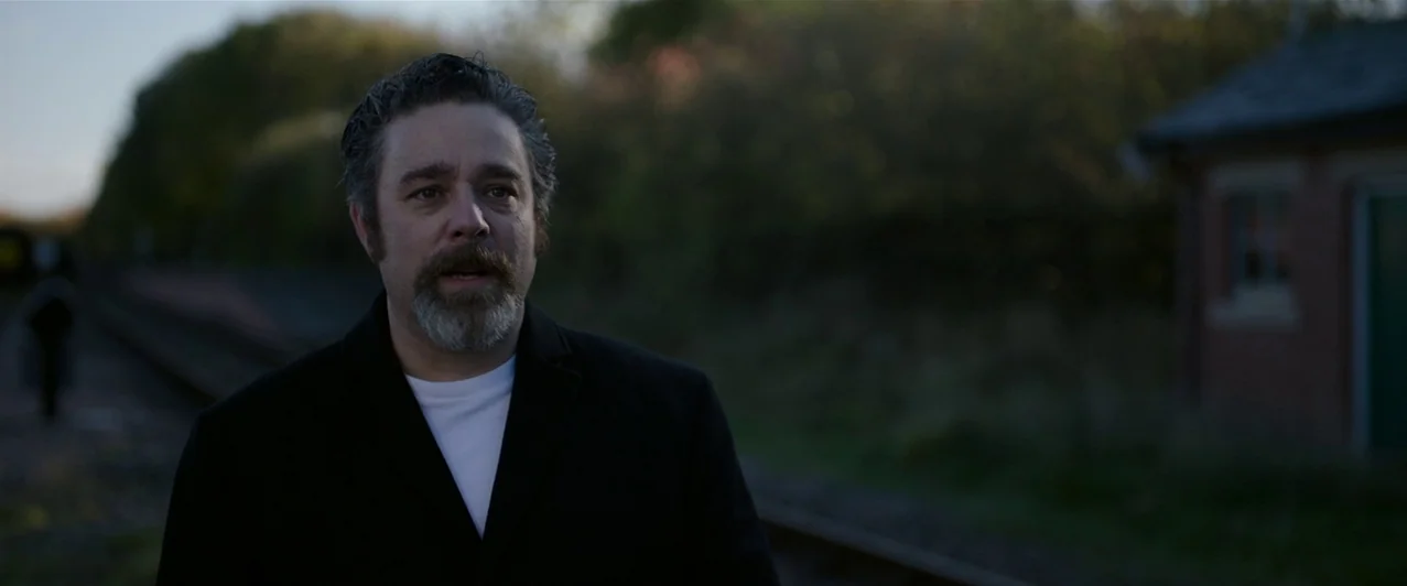 Ghost Stories (2017) movie still: medium — A quiet railway track area at dusk bordered by overgrown vegetation and a small building. A middle-aged…