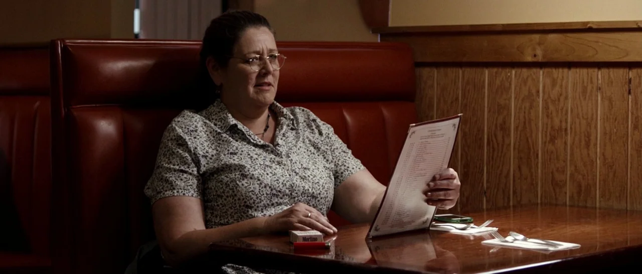 Cop Car (2015) movie still: medium — A woman sits in a red vinyl booth inside a diner with wood paneling. She holds a menu while looking off-camera…