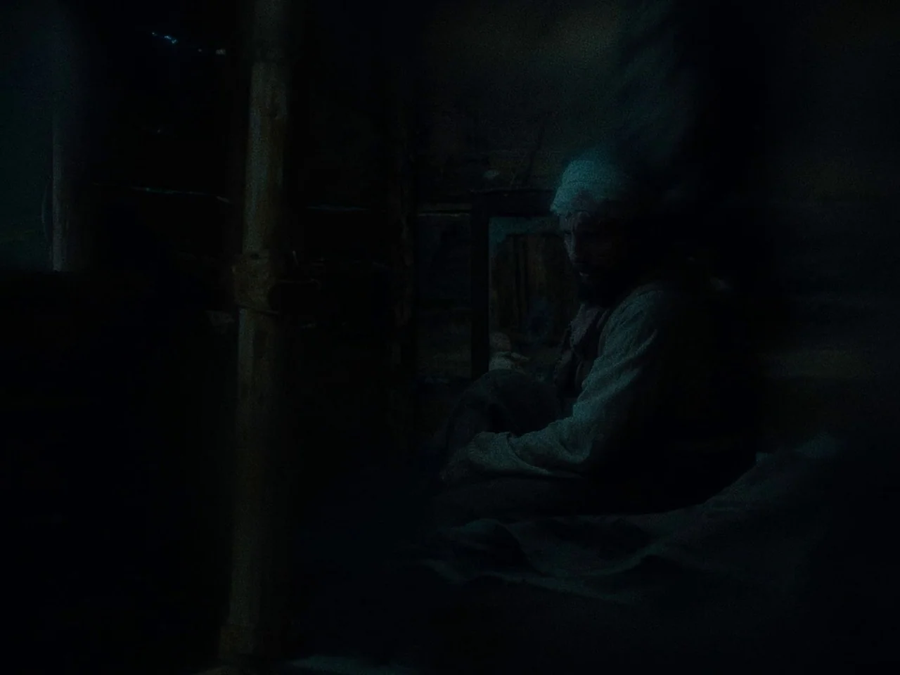 First Cow (2019) movie still: medium — A dark, dimly lit log cabin interior features rough wooden walls and shadows. A man sits alone in the low…
