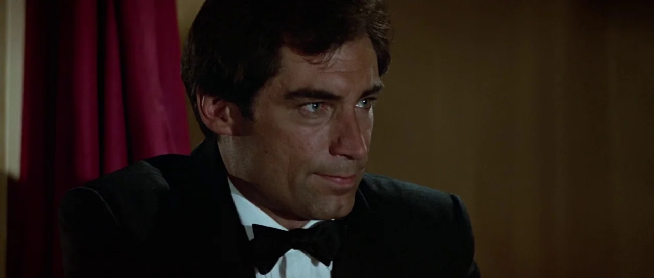 The Living Daylights (1987) movie still: closeup — A man in a tuxedo stands against a dark, featureless wall with red drapery. The man gazes toward…