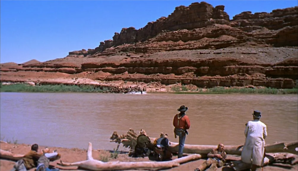 The Searchers movie still: wide — A group of armed men observe a group of figures crossing a brown river. High red rock cliffs form the background…