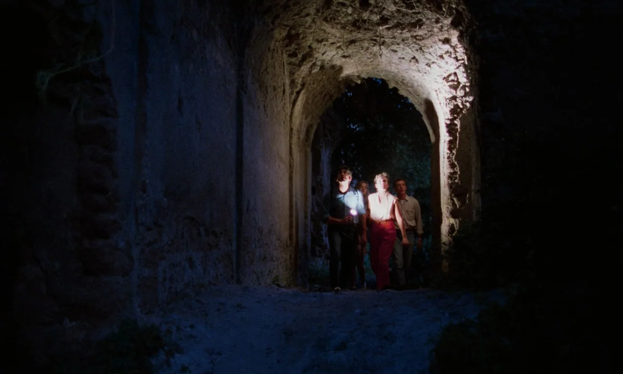 Demons (1985) movie still: fullbody — A group of people walks through a dark, weathered stone tunnel at night. They move cautiously while…