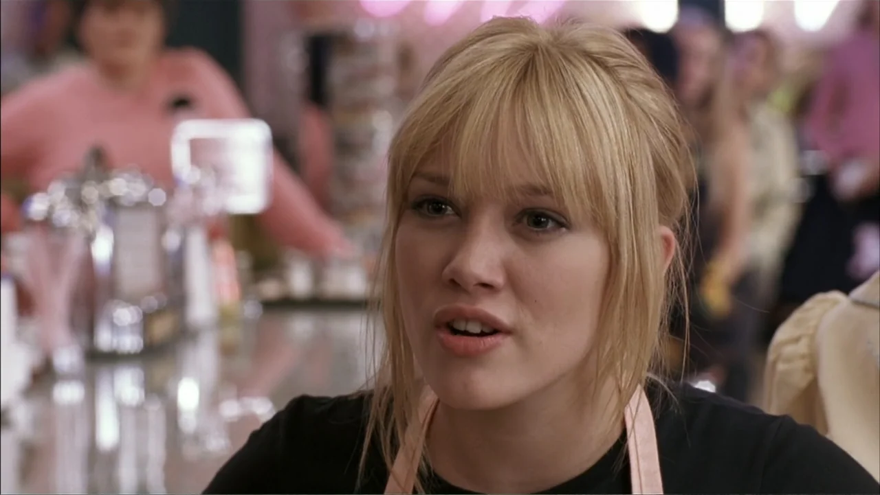 A Cinderella Story (2004) movie still: closeup — A brightly lit diner interior with a blurred background of patrons and equipment. A young woman in…