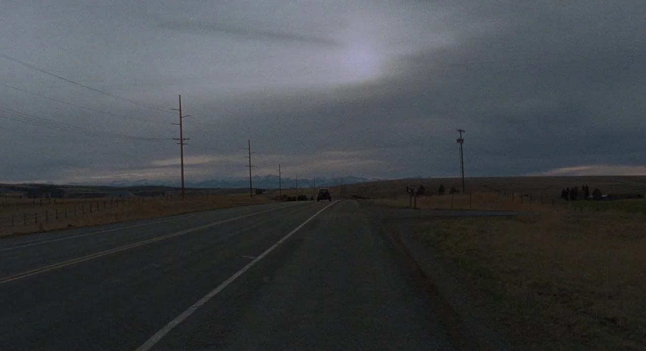 Certain Women (2016) movie still: wide — A long stretch of asphalt road cuts through a desolate, dry prairie landscape. A distant vehicle travels…