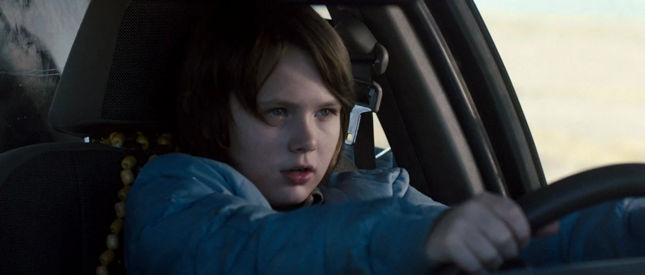 Cop Car (2015) movie still: closeup — A young boy sits in the driver seat of a vehicle during the daytime. He grips the steering wheel with both…