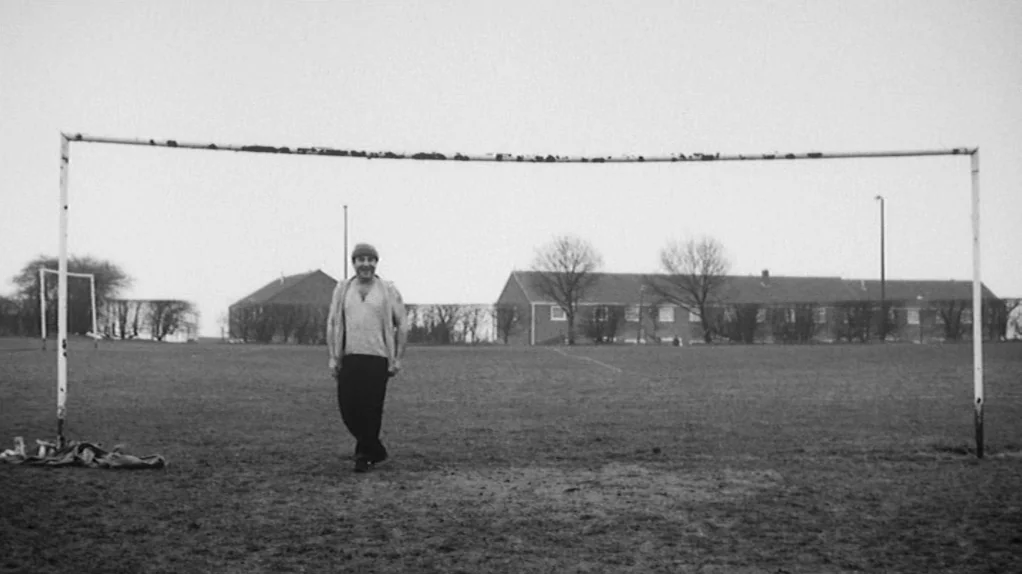 TwentyFourSeven (1997) movie still: fullbody — A bare grass sports field with a weathered metal goalpost in the foreground. A man stands still in…