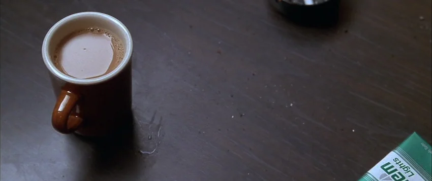Hard Eight (1996) movie still: closeup — A dark wooden surface holds a brown coffee mug and a pack of cigarettes. There is a small wet ring around…