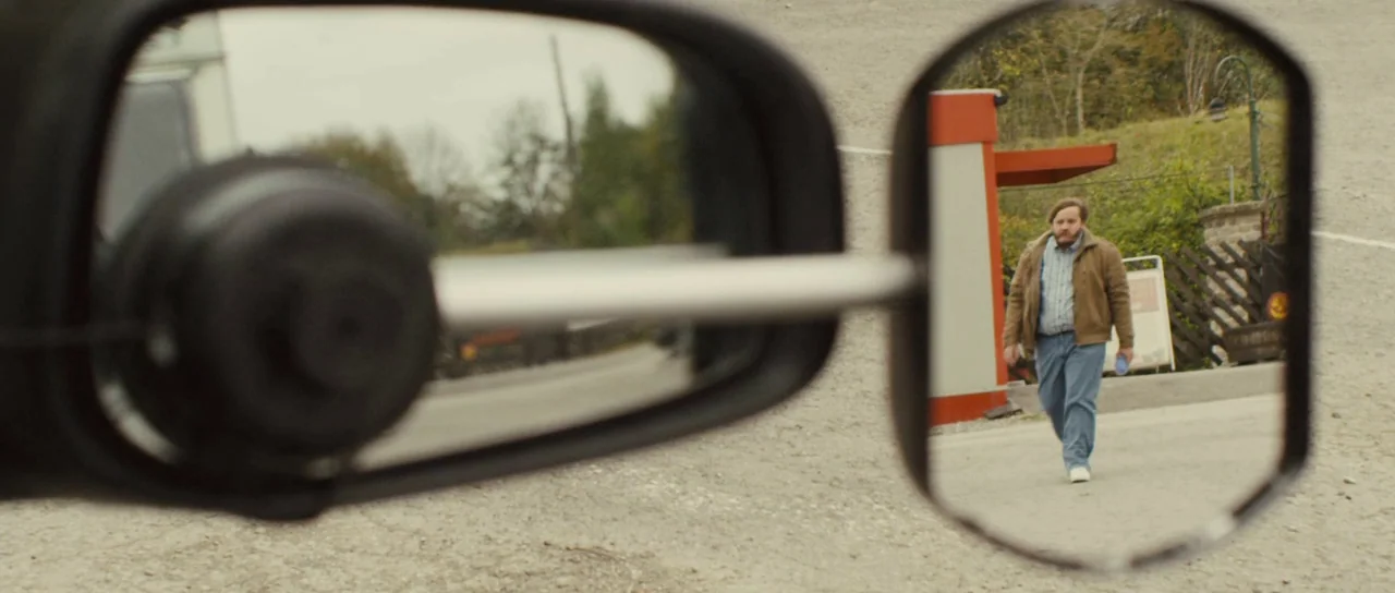 Sightseers (2012) movie still: closeup — A gravel parking area with a small red and white building structure. A man walks forward toward the camera…