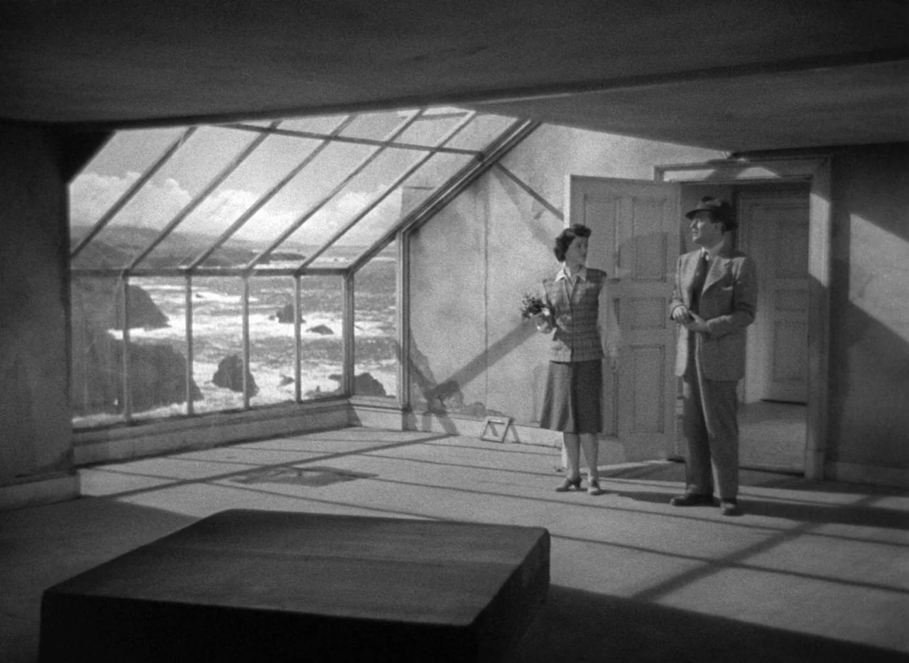 The Uninvited (1944) movie still: wide — An empty room with a large glass wall overlooking a rocky ocean coastline. A man and a woman are standing…