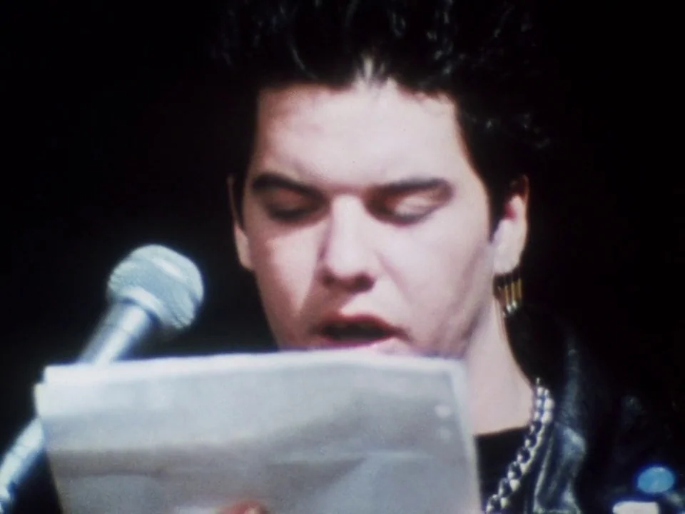Decline of Western Civilization (1981) movie still: closeup — A person stands on a dimly lit stage in front of a microphone. The subject reads text…