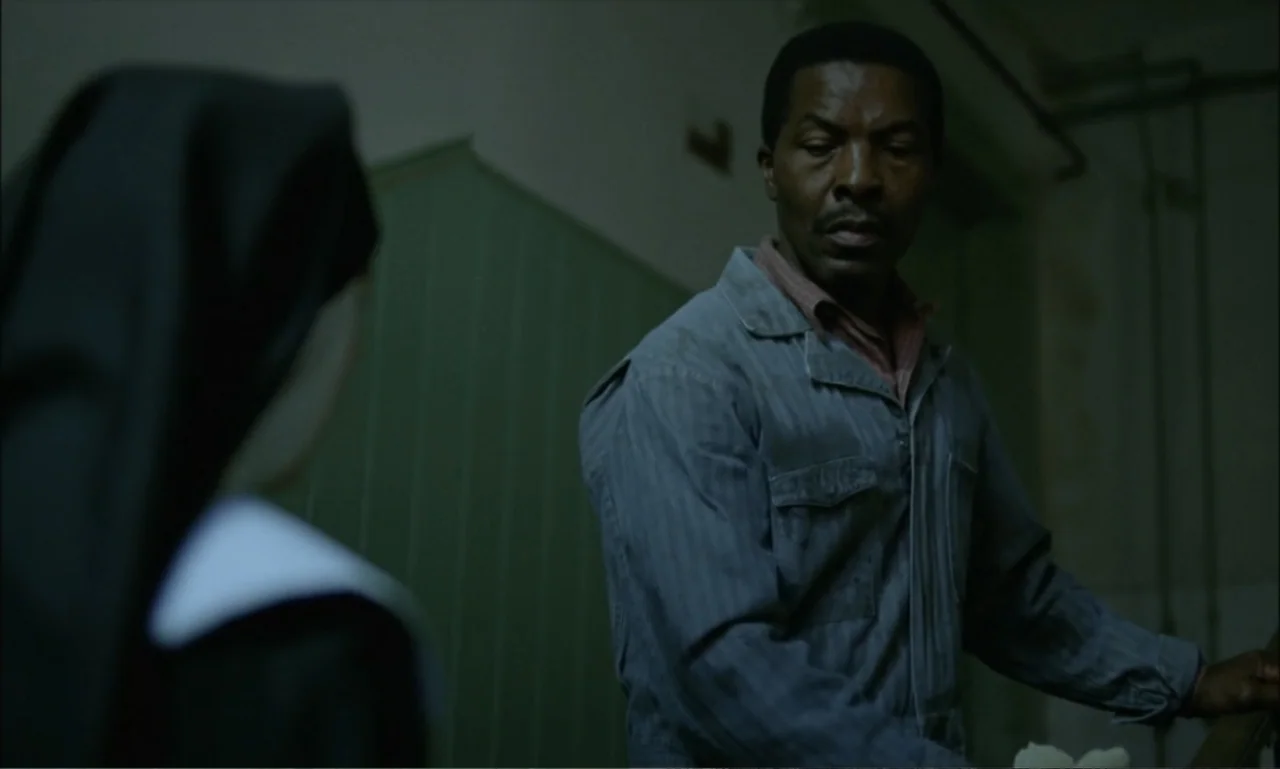 The Brutalist (2024) movie still: medium — An interior hallway with muted green walls and dimly lit architectural features. A man in a work jumpsuit…