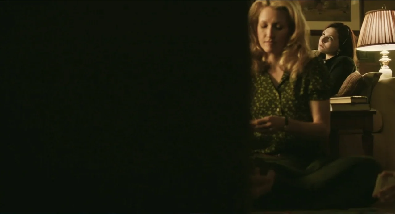 Haunter (2013) movie still: medium — A dimly lit room features a table lamp illuminating two women. One woman sits on the floor in the foreground…