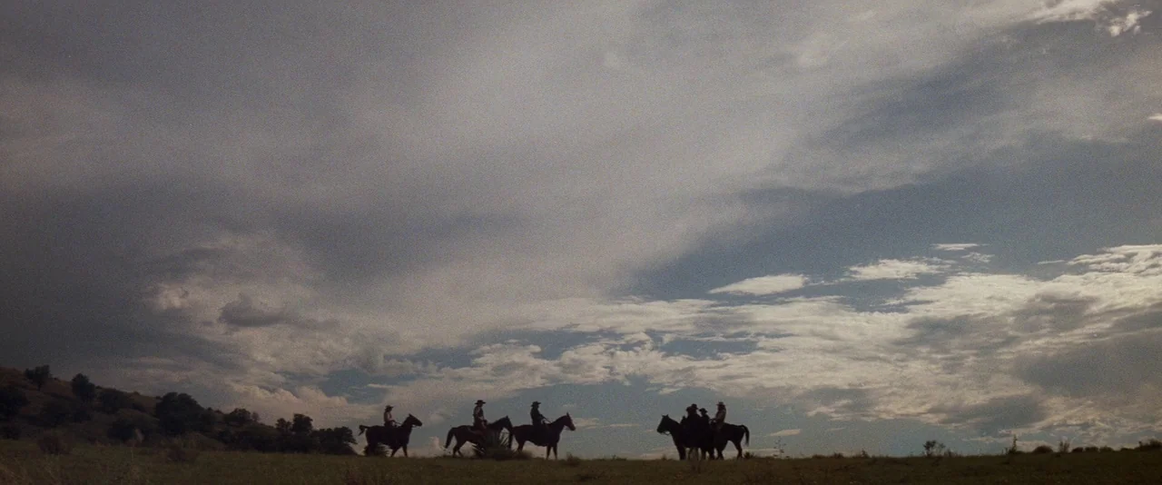 Tombstone (1993) movie still: wide — A vast, grassy landscape sits under a wide, cloudy sky in the afternoon light. A small group of people ride…