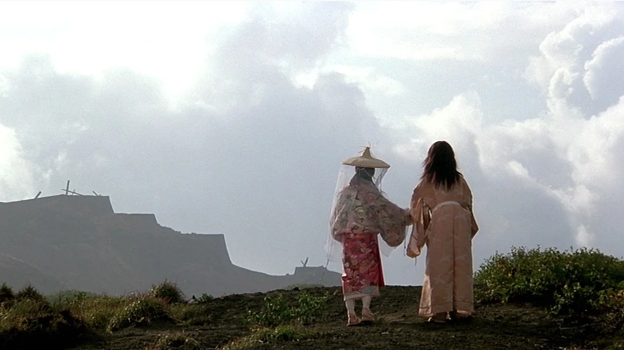 Ran (1985) movie still: wide — A grassy clifftop under a vast cloudy sky with distant arid hills and ridges. Two figures in traditional Japanese…