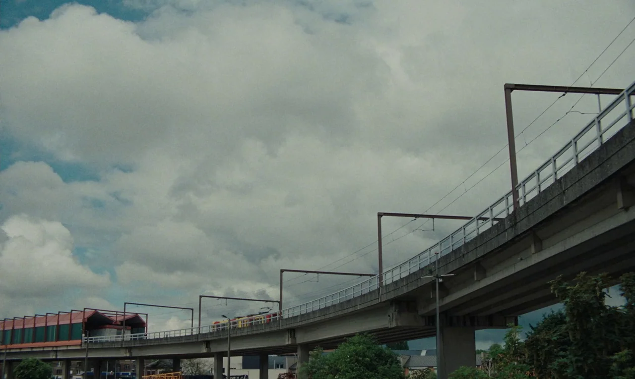 Maldoror (2024) movie still: establishing — An elevated concrete train viaduct stretches across the frame under a cloudy sky. A yellow and red train…