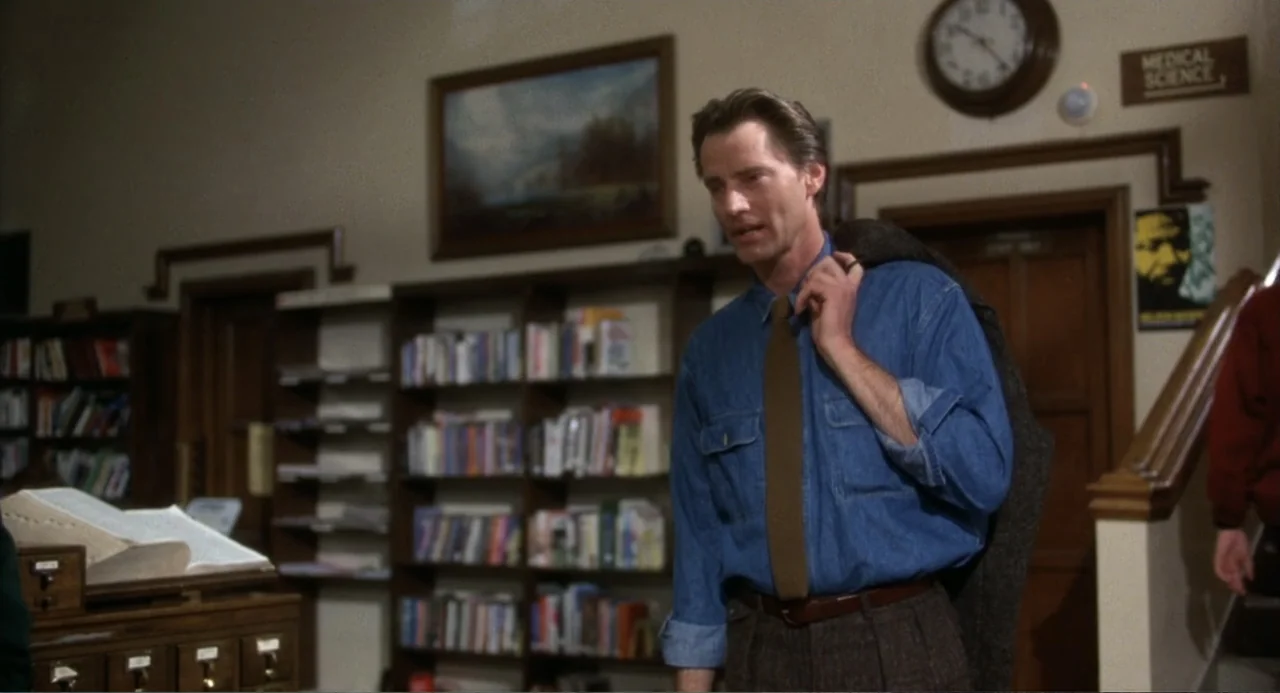 Baby Boom (1987) movie still: medium — A quiet library filled with bookshelves and a wall-mounted clock above a wooden door. A man in a blue denim…
