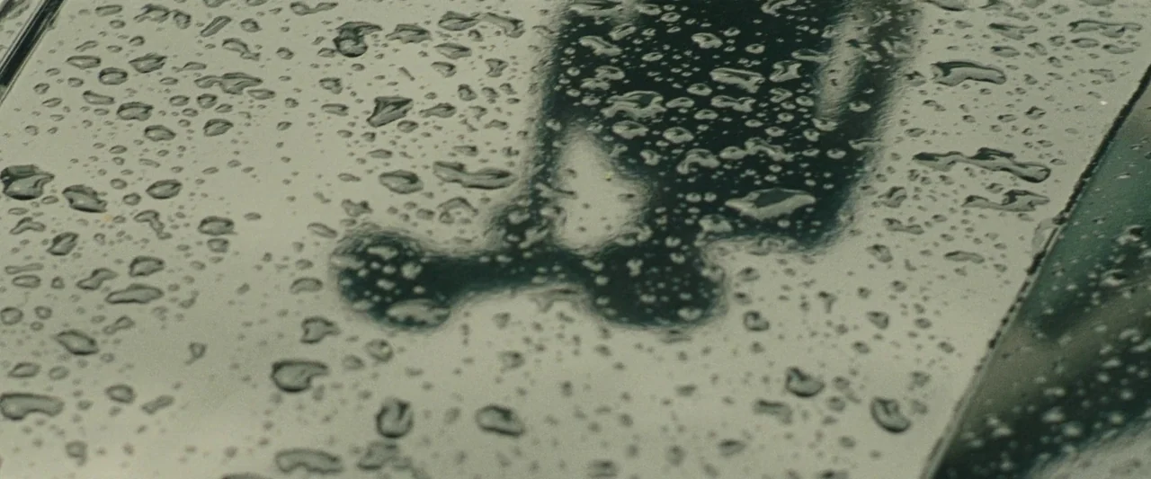 Mangrove (2020) movie still: closeup — A dark vehicle roof is covered in numerous water droplets after a rain shower. A blurry, dark silhouette of…