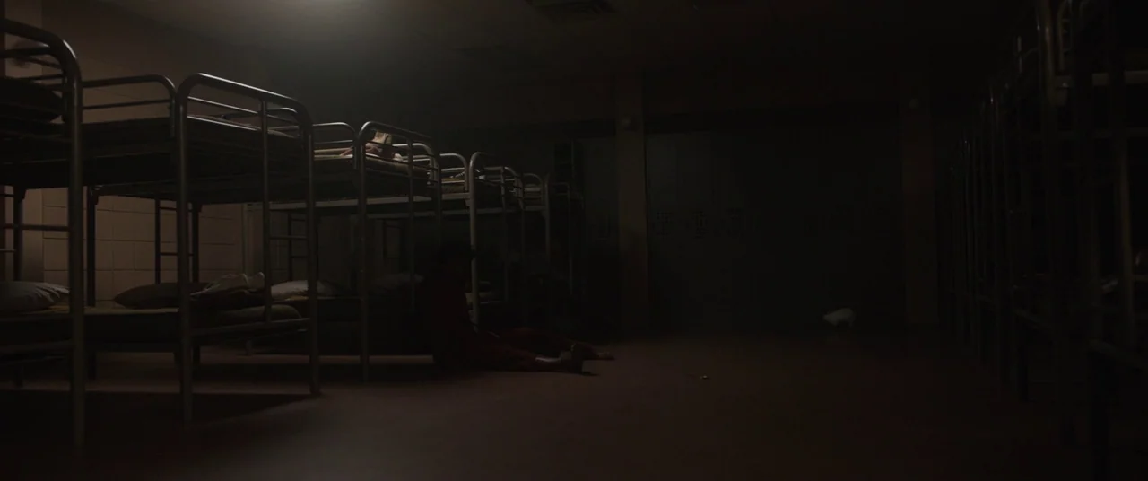 Us (2019) movie still: wide — A dimly lit industrial room contains multiple rows of metal bunk beds. A lone person in dark clothing sits on the…